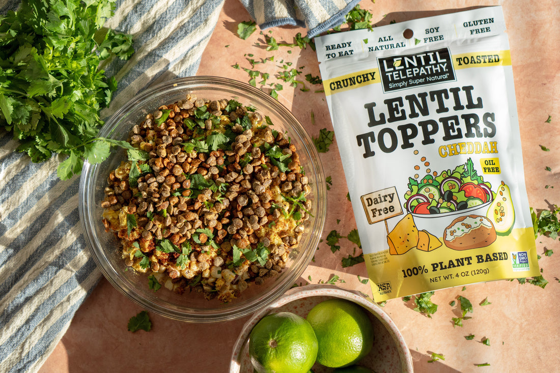 drilled elote corn dip topped with crunchy toasted lentils from Lentil Telepathy displayed in a bowl on a table alongside cilantro and lime garnish and a farm country striped dishtowel